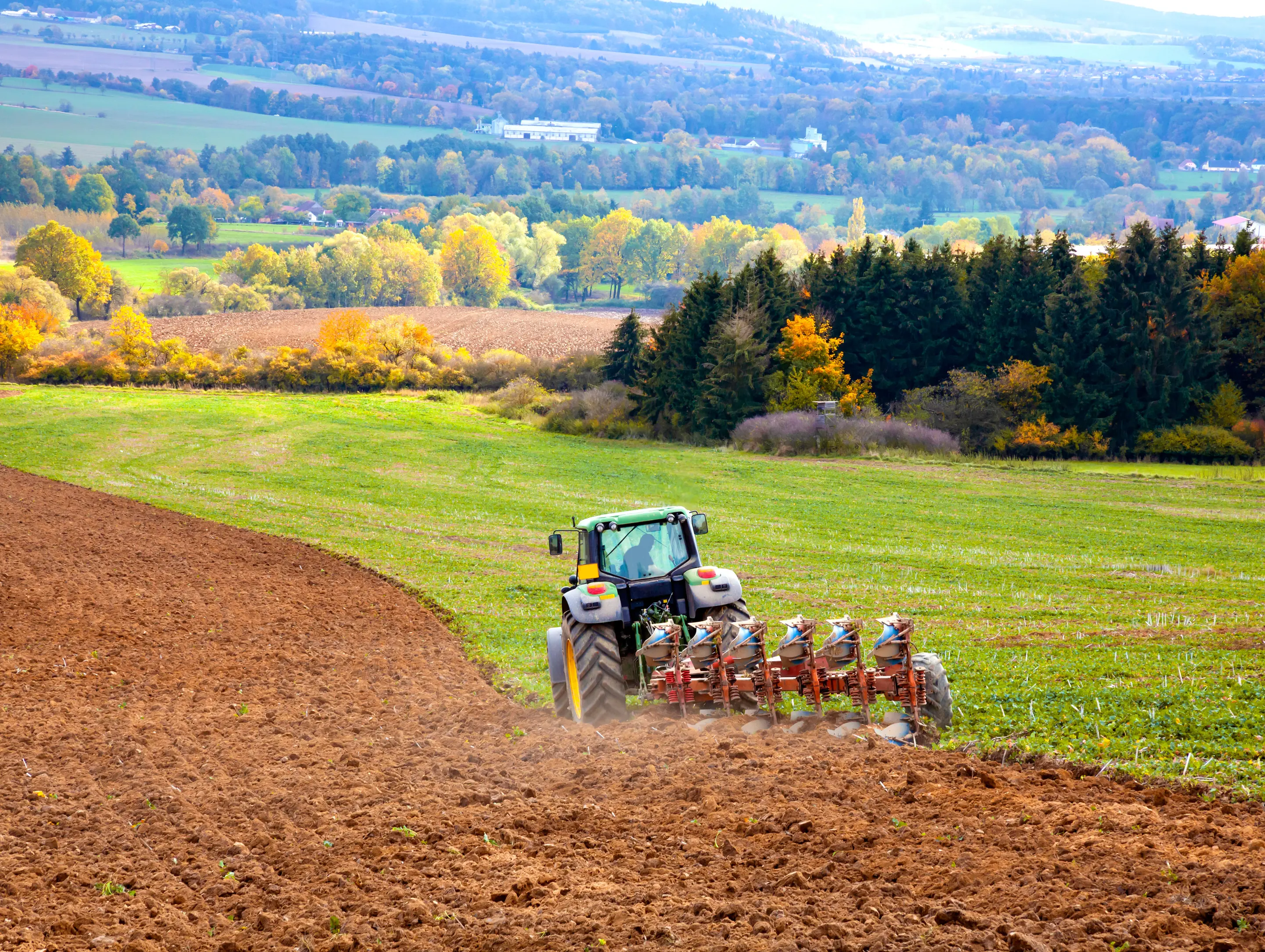 Zelenomodrý traktor s pripojeným viacradovým pluhom vykonáva hlbokú orbu na mierne svahovitom poli. Na ľavej strane je čerstvo vyoraná, hnedá a kyprá zemina, kým vpravo a pred traktorom sa nachádza zelené pole so zvyškami vegetácie. V pozadí je pestrá vidiecka krajina s lesíkmi, poliami s jesenným sfarbením stromov, dedinou a vzdialenými kopcami pod svetlou oblohou.